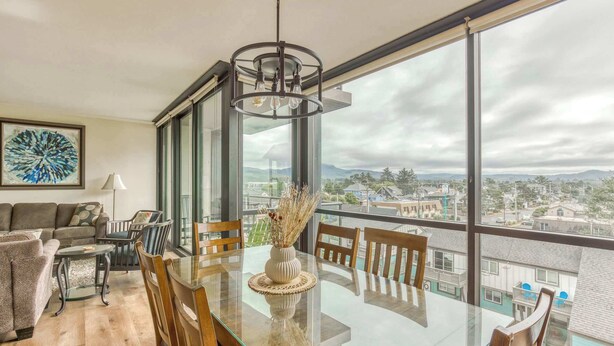 A spacious living room with a glass dining table, comfortable seating, a large window with a city view, and a modern chandelier.