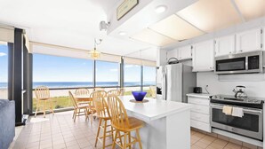 A modern kitchen with white cabinets, stainless steel appliances, and a large island. There is a dining area with wooden chairs and a view of the ocean through large windows. - Sand & Sea (402) Water's Edge Panoramic Views (Seaside)