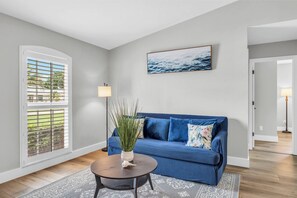 Ein Wohnzimmer mit einem blauen Sofa, einem runden Couchtisch, einer Pflanzvase, einem gerahmten Bild an der Wand und einem Fenster mit Jalousien.
