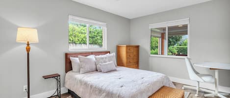 A bedroom with a bed, bedside lamp, dresser, chair, and a window with a view of greenery.