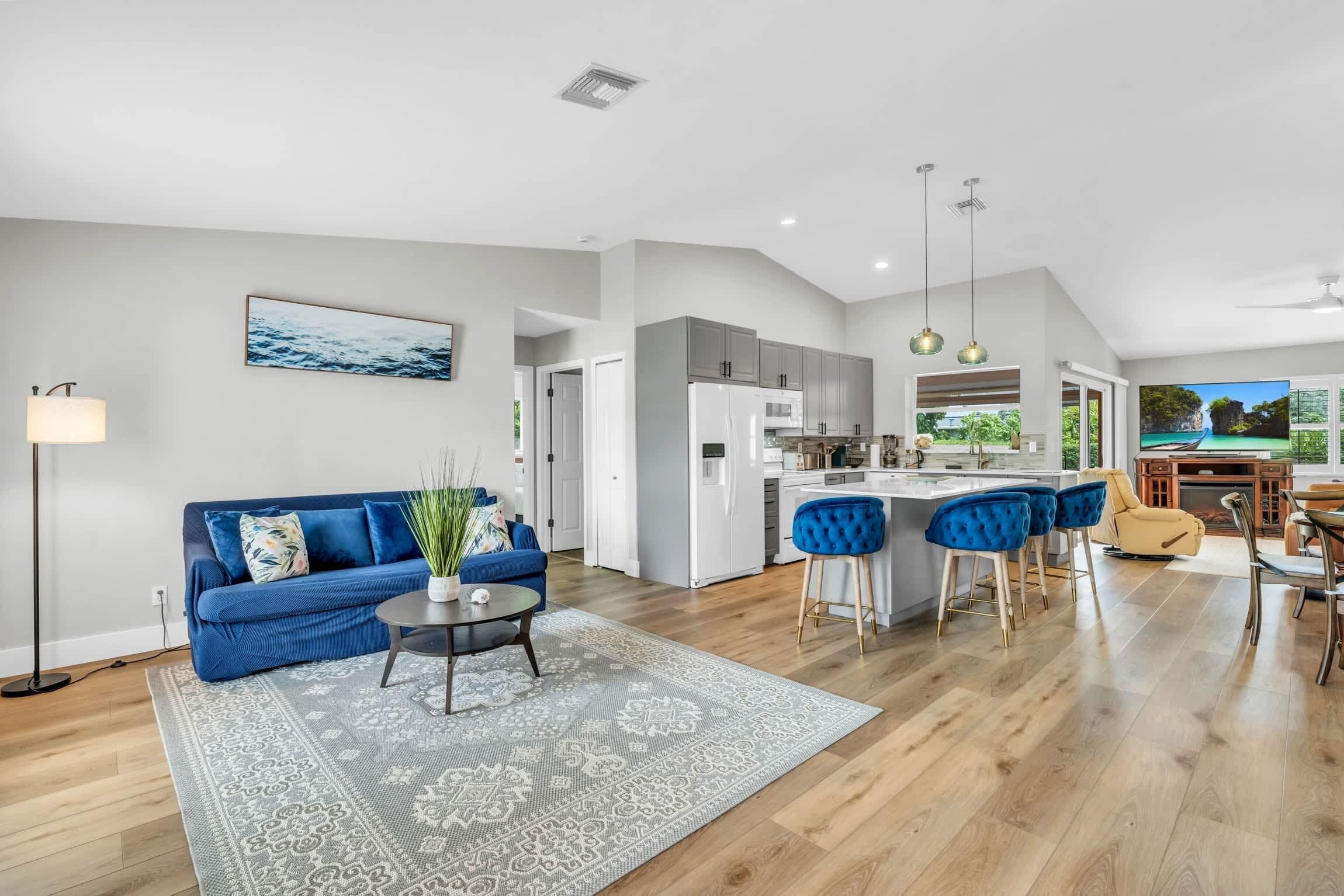 A modern living space with a blue sofa, a dining area with bar stools, a kitchen, and a large window with a view.