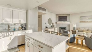 A modern kitchen with white cabinetry and a marble island, connected to a dining area with a wooden table and chairs. - Sand & Sea (508) Salt Life - Lux Beachfront w Pool (Seaside)