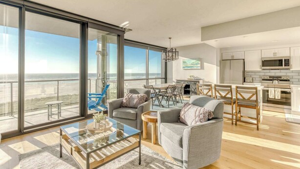 A modern living room with a glass coffee table, grey sofas, and a dining area with wooden chairs. A kitchen with white cabinets and a large window is visible in the background. - Sand & Sea (508) Salt Life - Lux Beachfront w Pool (Seaside)
