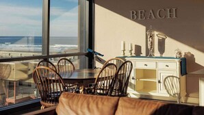 A dining area with a view of the beach, a table, chairs, and a cabinet with decorative items. - Sand & Sea (618) Seaside Panorama -Cozy Beachfront (Seaside)