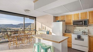 A modern kitchen with wooden cabinets, a white refrigerator, a microwave, and a stove. There is a dining area with a table and chairs, and a view of the mountains through large windows. - Sand & Sea (618) Seaside Panorama -Cozy Beachfront (Seaside)