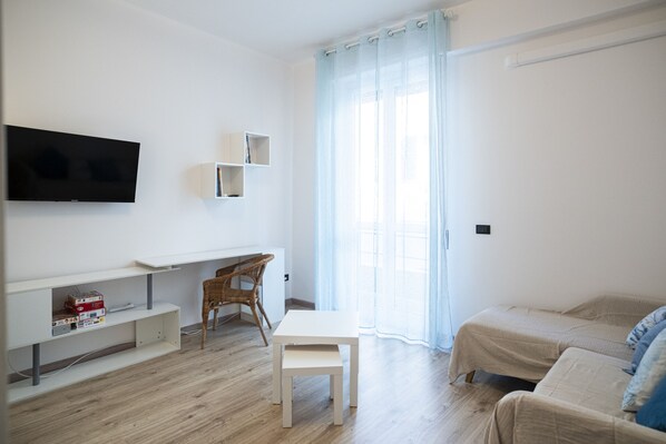 A modern, minimalist room with a flat-screen TV mounted on the wall, a small white desk with a chair, a bed with a beige cover, and a large window with sheer curtains. - Lupo di Mare by Wonderful Italy (Savona)