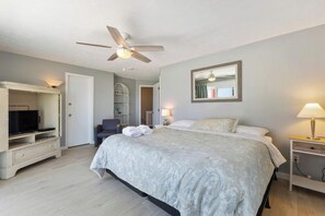 Une chambre à coucher comprenant un lit, un téléviseur, un ventilateur de plafond, une chaise, une table de chevet et une commode.