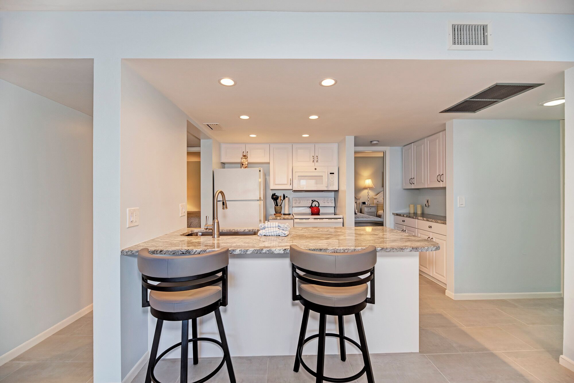 A modern kitchen with a central island, two bar stools, white cabinetry, and a microwave.