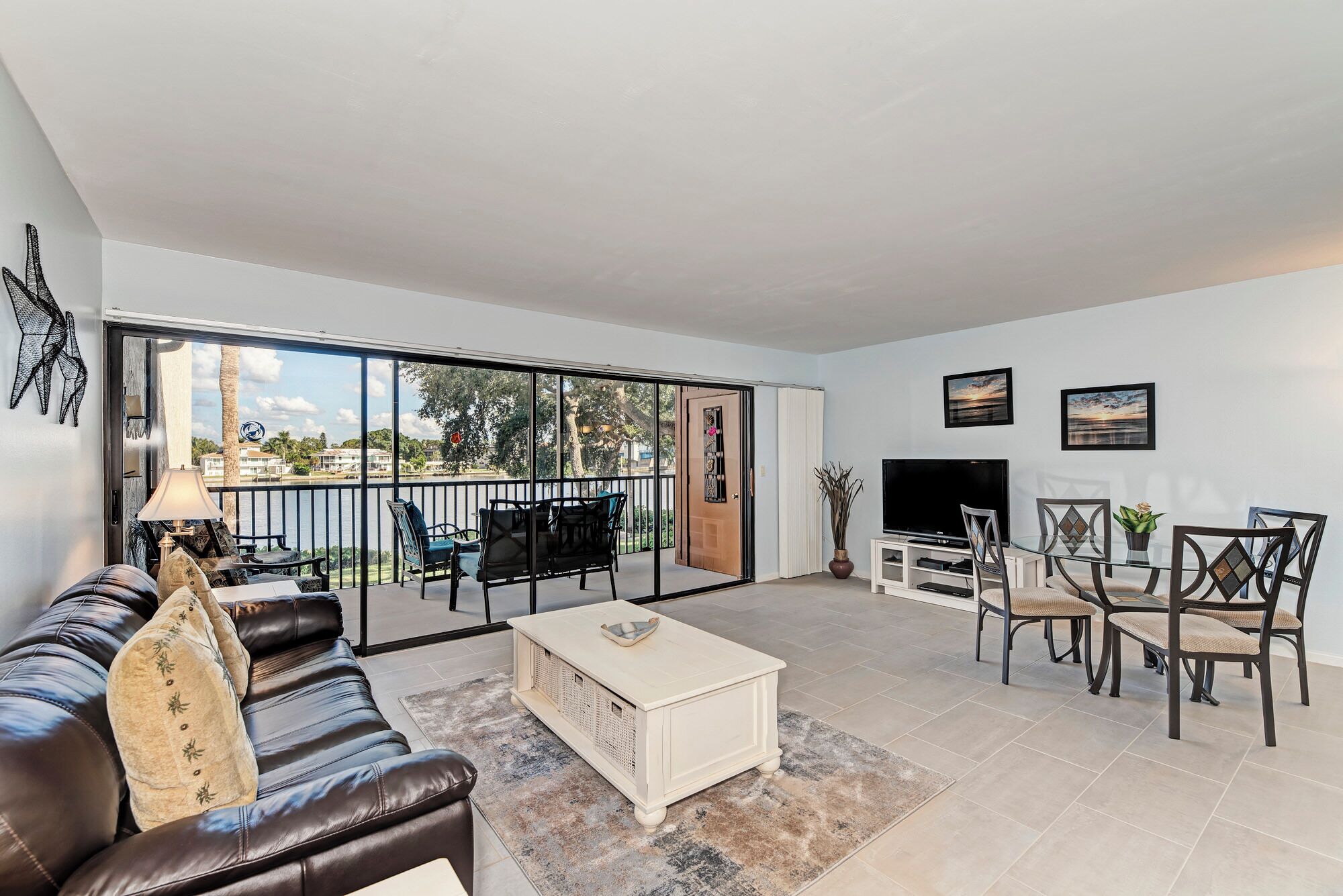 A spacious living room with a large sliding glass door leading to a balcony, a comfortable sofa, a coffee table, a TV, and a dining area with chairs.