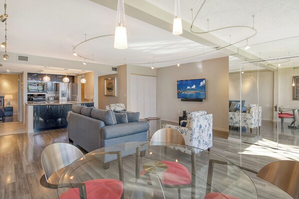 A modern living room with a glass dining table, sofas, and a kitchen in the background. - Siesta Key Condo, Private Beach Access & Water Views - Midnight Cove II 122F (Siesta Key)