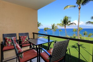 A balcony with a table and chairs, overlooking a beach with palm trees and the ocean. - Kihei Surfside, #511 1 Bedroom Condo by RedAwning (Kihei)