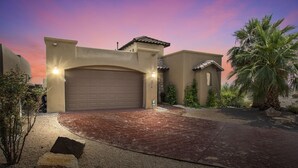 Een woonhuis met garage, oprit en een verzorgde tuin met palmbomen en struiken.