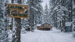 Un panneau en bois sur lequel on peut lire « ESCAPADE EN ALPES » et une cabane entourée d’arbres couverts de neige.