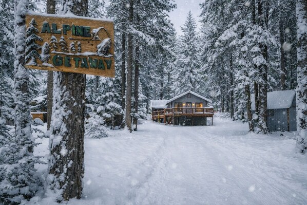 Un segnale di legno con la scritta 'ALPINE GETAWAY' e una baita circondata da alberi innevati.