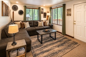 A living room with a sectional sofa, a coffee table, a lamp, and a view of the outdoors through large windows. - Alpine Getaway 2 Bedroom Home by RedAwning (Leavenworth)