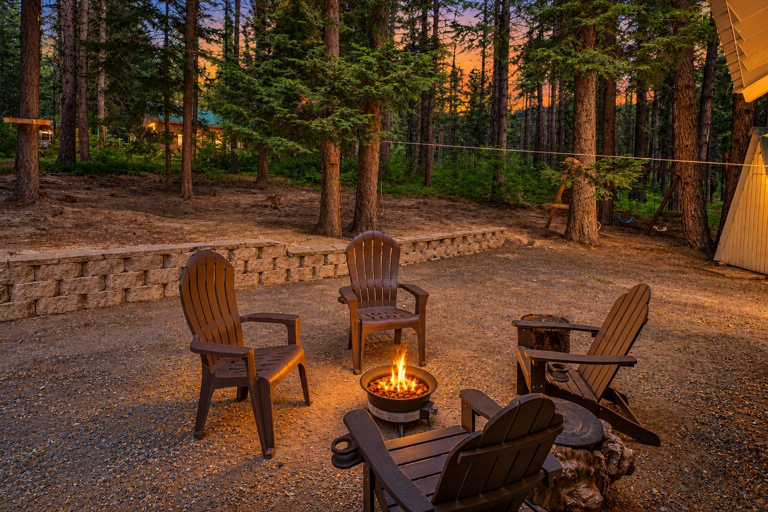 A fire pit with four Adirondack chairs arranged around it, set in a forested area with a stone wall and a wooden structure in the background.