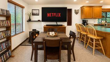 A dining area with a TV displaying 'NETFLIX,' a fireplace, wooden chairs, and a bookshelf filled with DVDs.