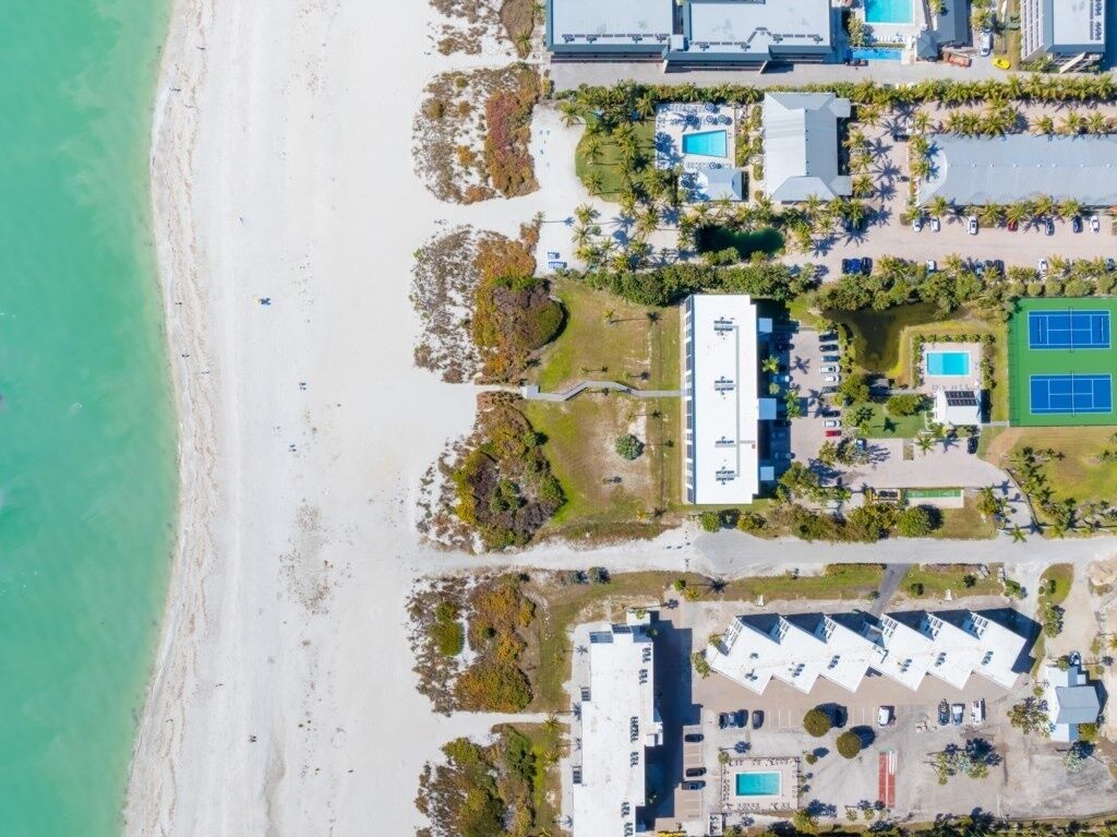 Vue aérienne d’une propriété située sur la côte, avec une plage, des piscines, des courts de tennis et un grand bâtiment doté de nombreux espaces de stationnement.