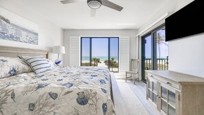A bedroom with a bed, a desk, a chair, a TV, and a large window with a view of the beach. - Pelicans Roost 203 2 Bedroom Condo by RedAwning (Sanibel)