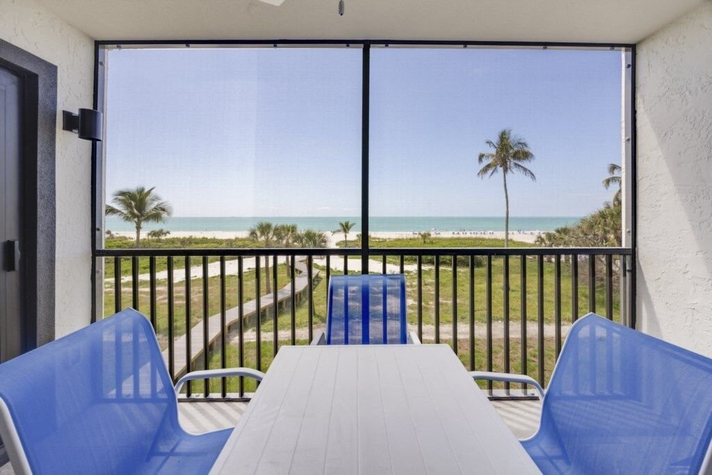 A balcony with a white table and blue chairs, overlooking a beach with palm trees and a clear sky.