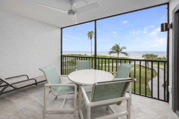 A balcony with a white table and chairs, a ceiling fan, and a view of the beach. - Pelicans Roost 204 2 Bedroom Condo by RedAwning (Sanibel)