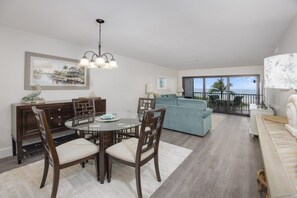 A modern living room with a round dining table, wooden chairs, a blue sofa, and a balcony with a view of the ocean. - Pelicans Roost 204 2 Bedroom Condo by RedAwning (Sanibel)