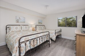 A bedroom with two beds, a wooden dresser, and a window with a view of palm trees. - Pelicans Roost 204 2 Bedroom Condo by RedAwning (Sanibel)