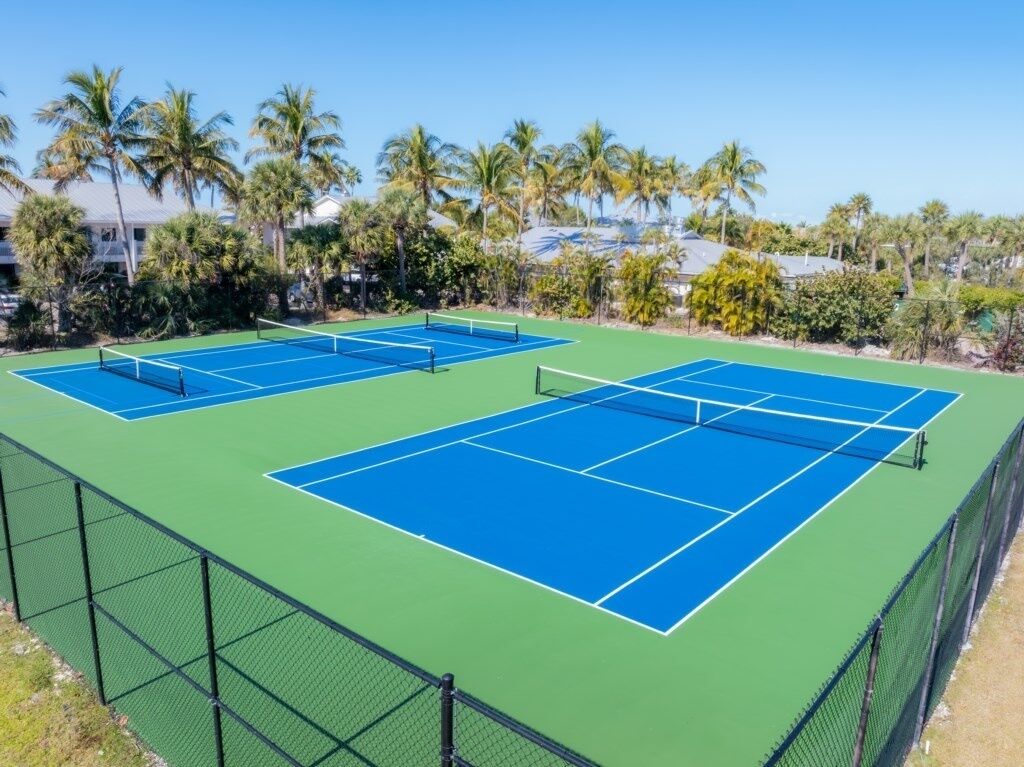 A tennis court with a green surface and blue lines, surrounded by a fence and palm trees.