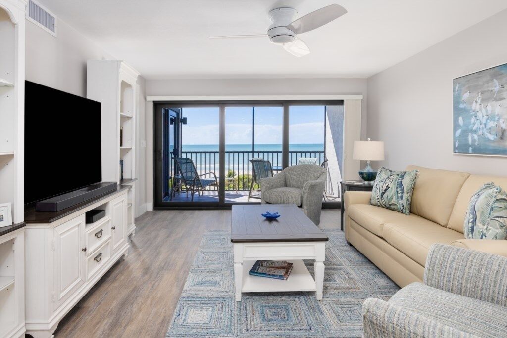 A living room with a TV, sofa, coffee table, and chairs. There is a balcony with a view of the ocean.