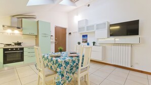 Cocina compacta con estufa, horno y gabinetes blancos. Hay una mesa de comedor con sillas y un televisor montado en la pared.