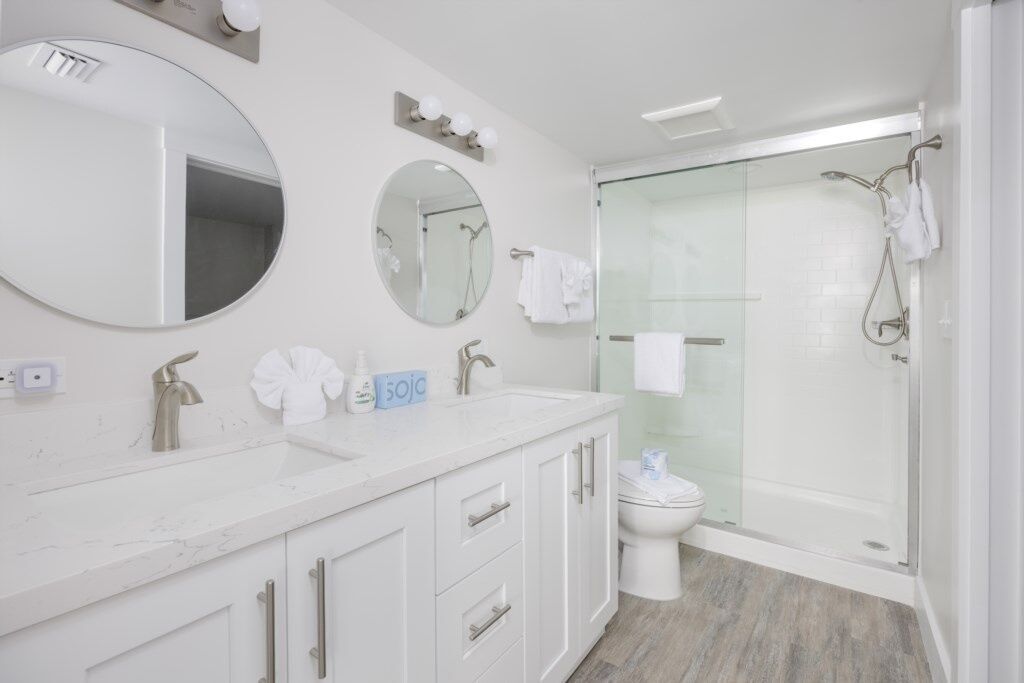 A modern bathroom with a white vanity, two sinks, a toilet, and a glass-enclosed shower.