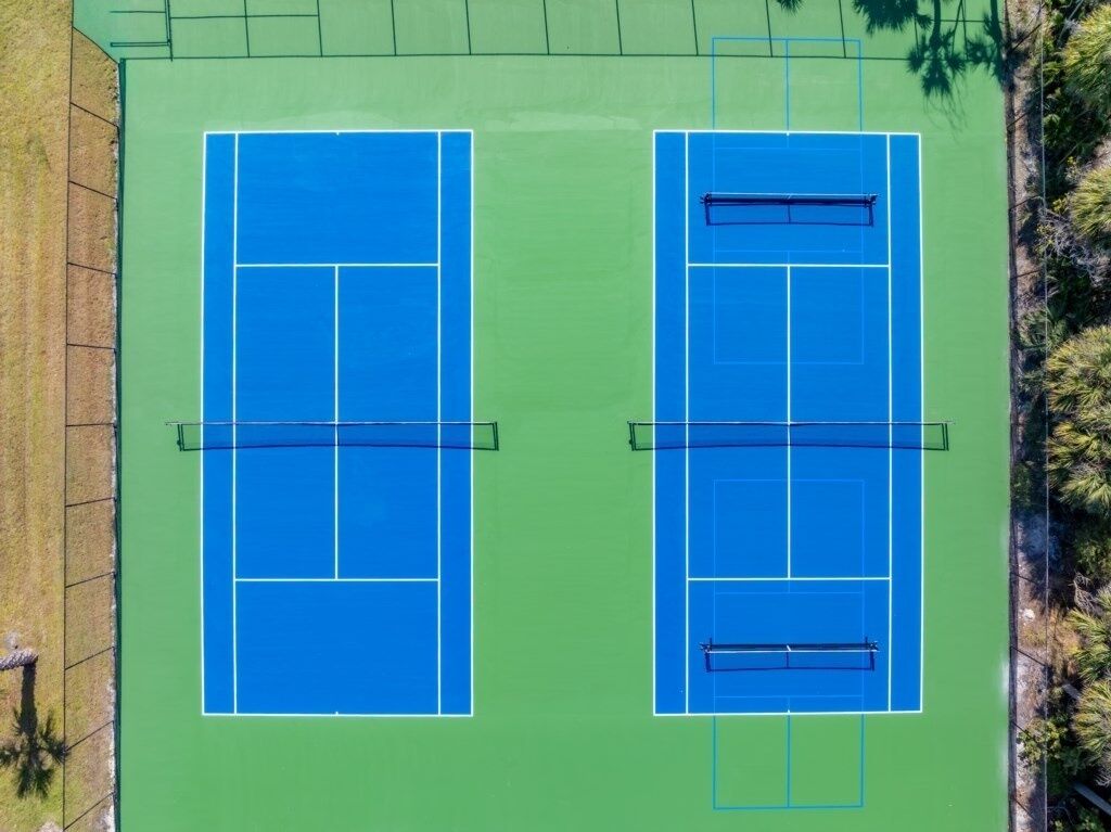 Zwei angrenzende Tennisplätze mit Netzmarkierungen, umgeben von Grünflächen und Bäumen.