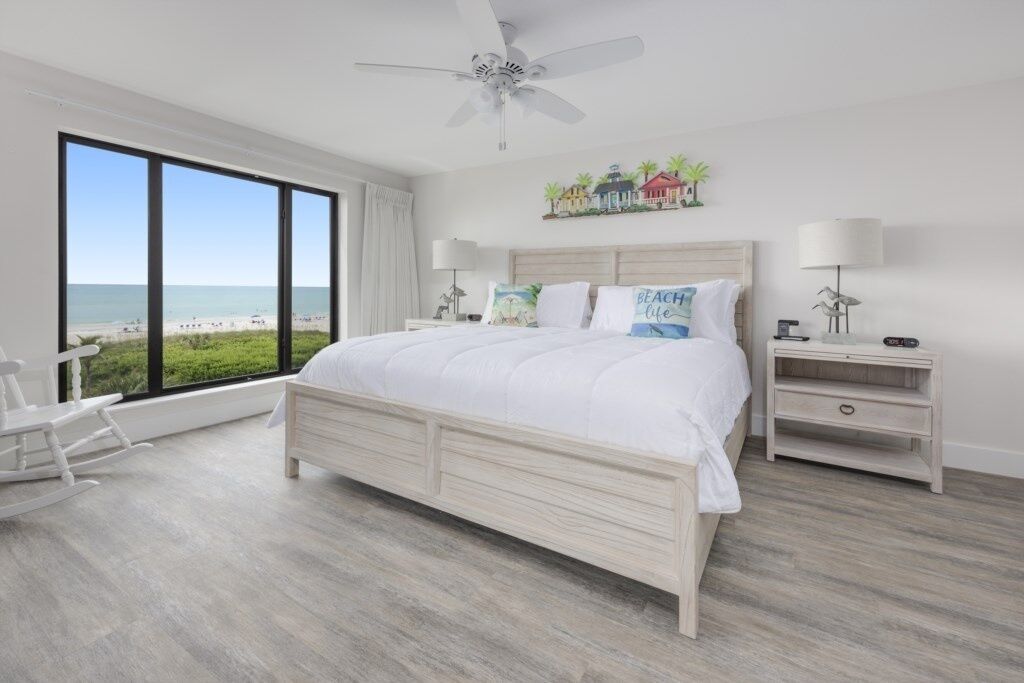 A bedroom with a large bed, a ceiling fan, a view of the beach, and a rocking chair.