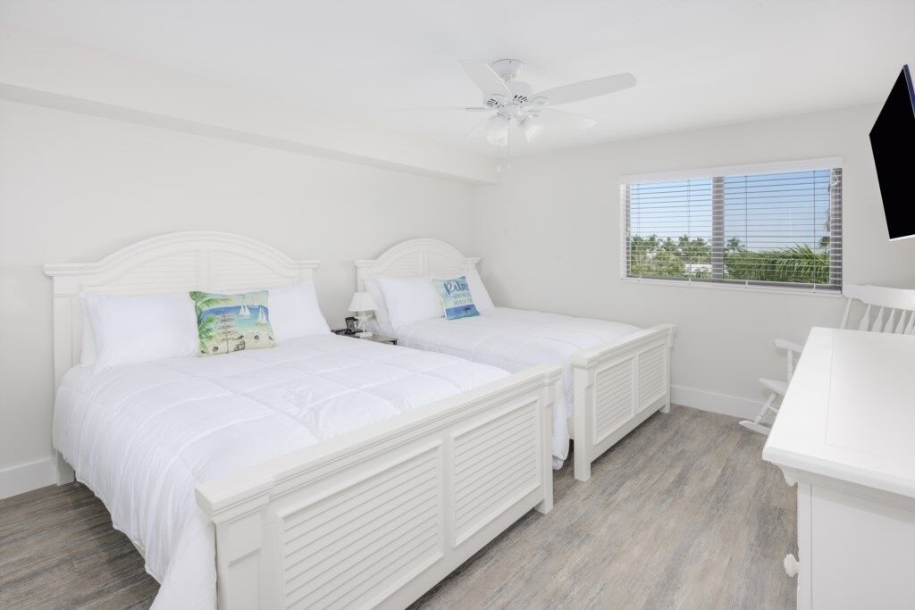 A bedroom with two white beds, a ceiling fan, a window with a view, and a white dresser.