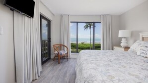 A hotel room with a large bed, a chair, a TV, and a view of the beach. - Pelicans Roost 202 2 Bedroom Condo by RedAwning (Sanibel)