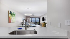 A modern kitchen with a stainless steel sink, white countertops, and grey cabinets. There is a large window providing a view of the outdoors. - Pelicans Roost 202 2 Bedroom Condo by RedAwning (Sanibel)