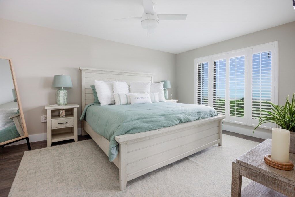 A bedroom with a white bed, two bedside tables, a ceiling fan, and a large window with blinds.