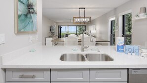 A modern kitchen with a double sink, white cabinetry, and a view to the dining area. - Pelicans Roost 101 2 Bedroom Condo by RedAwning (Sanibel)