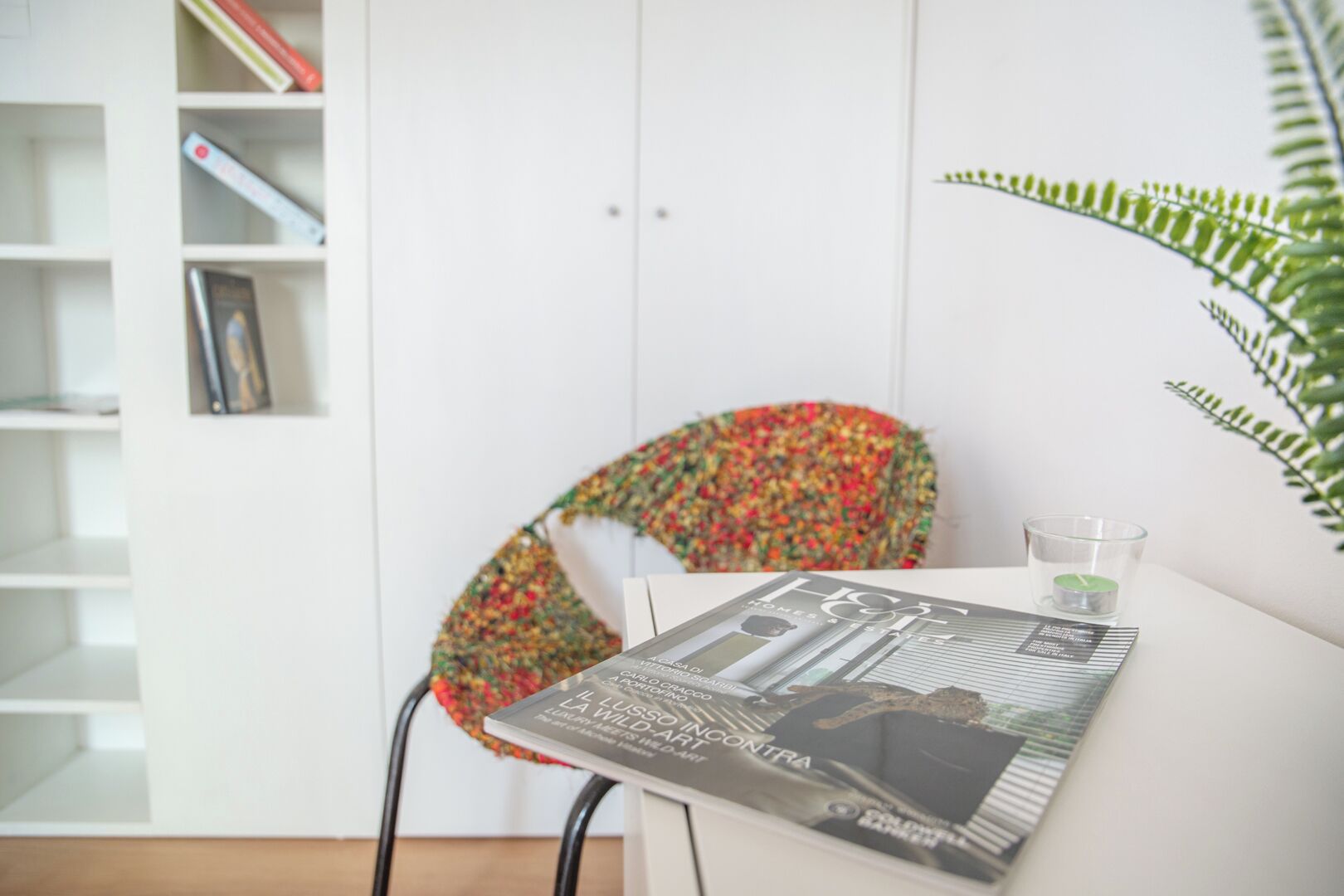 Une pièce avec une table blanche, une chaise dont le dossier est fait d’un tissu coloré, un verre d’eau et une plante.