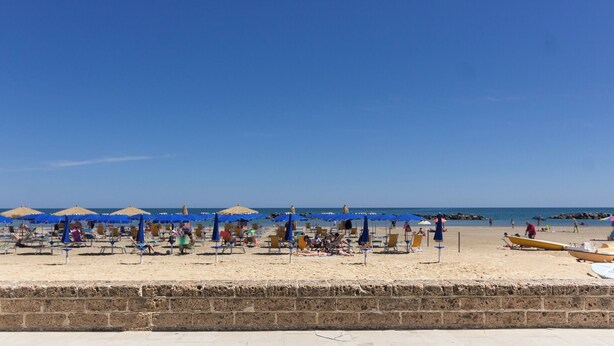 Une plage bordée de rangées de chaises longues et de parasols, un mur de pierre au premier plan et l’océan en arrière-plan.