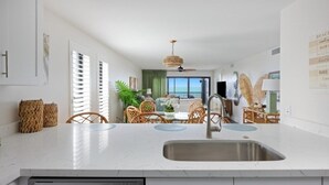 A modern kitchen with a built-in dishwasher, a dining area with wicker chairs, and a view of the ocean. - Pelicans Roost 207 2 Bedroom Condo by RedAwning (Sanibel)