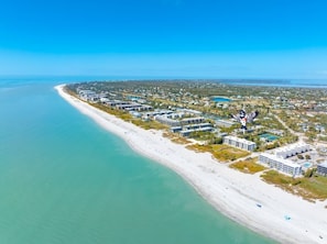 Une vue aérienne d’un hôtel situé sur la côte, composé de plusieurs bâtiments le long de la plage, dont une zone avec piscine et un mât portant un drapeau.