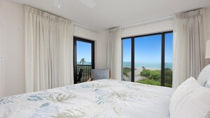 A bedroom with a large bed, two windows with ocean views, and a balcony with a chair. - Pelicans Roost 207 2 Bedroom Condo by RedAwning (Sanibel)