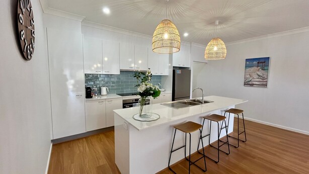 A modern kitchen with white cabinets, a central island with stools, a sink, and two pendant lights. - Jo Sea by Book Jervis Bay - Luxury in Vincentia with Ocean Views (Vincentia)