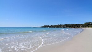 A beach with clear blue water, a sandy shore, and a line of trees in the background. - Jo Sea by Book Jervis Bay - Luxury in Vincentia with Ocean Views (Vincentia)
