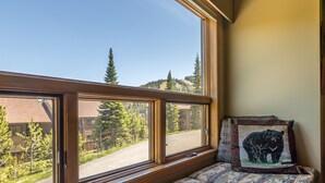 A room with a large window overlooking a road and trees, a bed with patterned pillows, and a bear-themed pillow.
