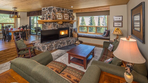 A cozy living room with a stone fireplace, comfortable seating, and large windows overlooking a forested area.