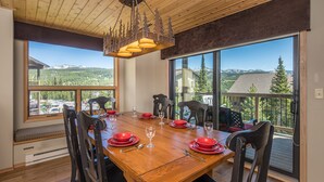 A dining area with a wooden table set for four, large windows offering mountain views, and a ceiling with a tree design.