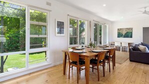 Un coin repas avec une table et des chaises en bois, de grandes fenêtres donnant sur un paysage verdoyant, un canapé gris et un ventilateur de plafond.