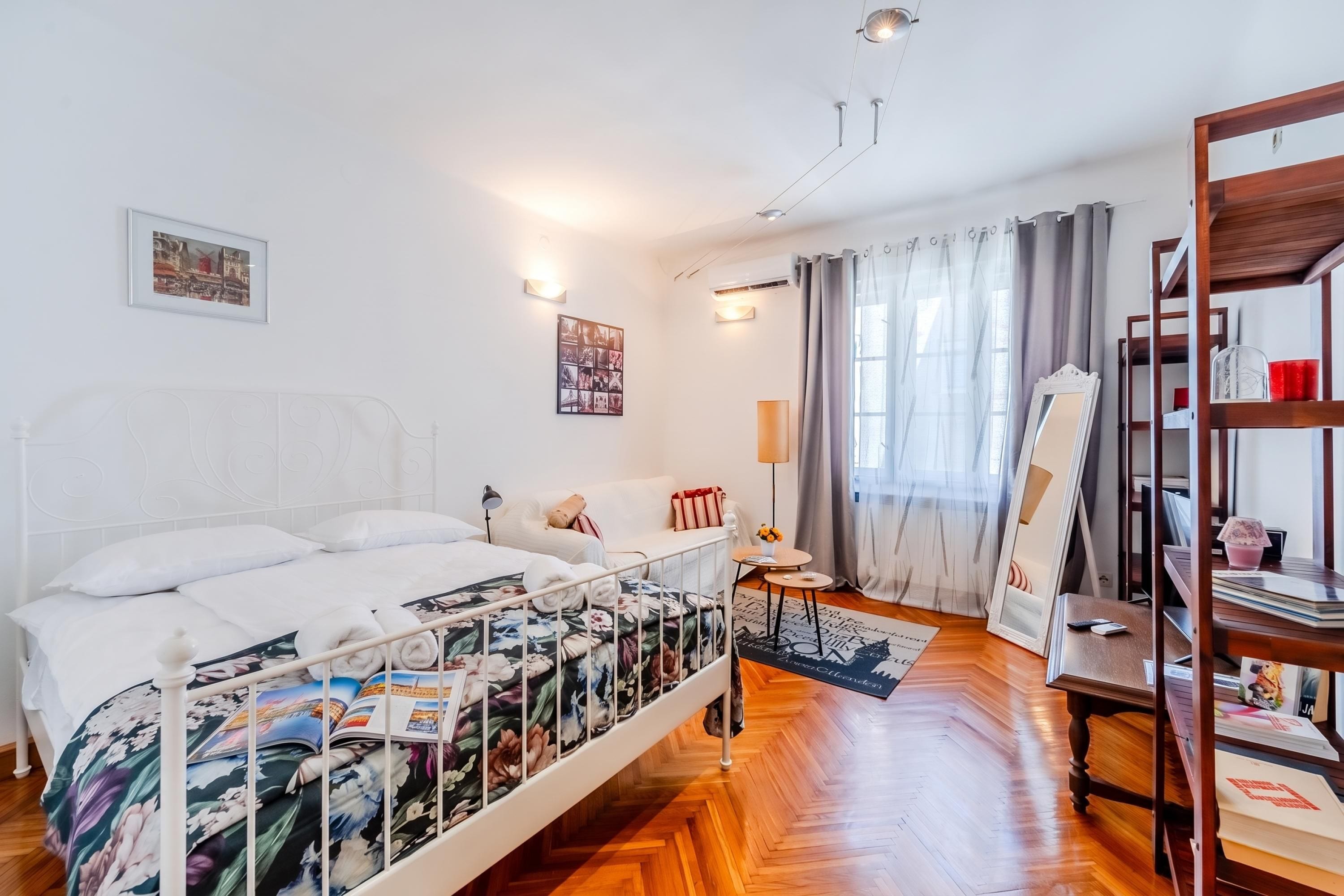Une chambre d’hôtel avec un lit, une table de chevet, une lampe, un miroir, une étagère avec des livres et une fenêtre avec des rideaux.